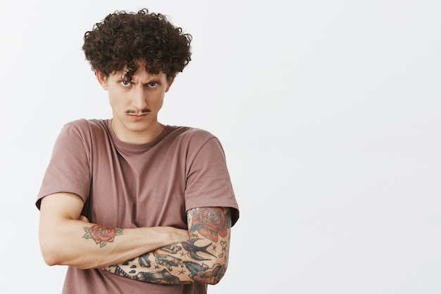 Curly-haired young man with tattoos folding arms and looking skeptical