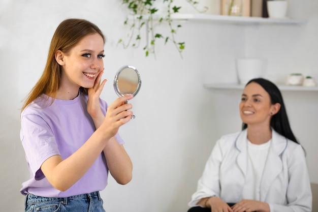 A woman smiling while looking at a mirror, with another woman in a white coat observing in the background.