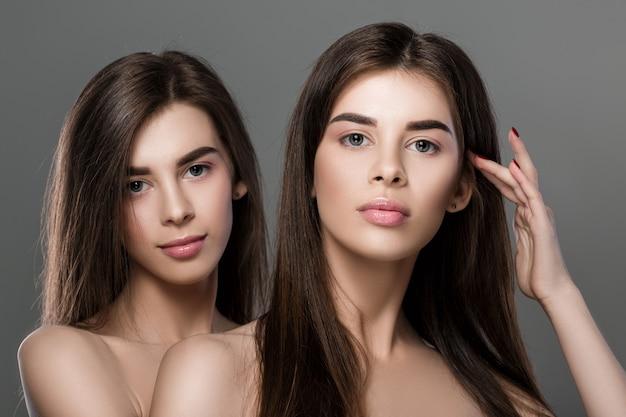 Two young women with flawless skin and dark hair posing against a gray background