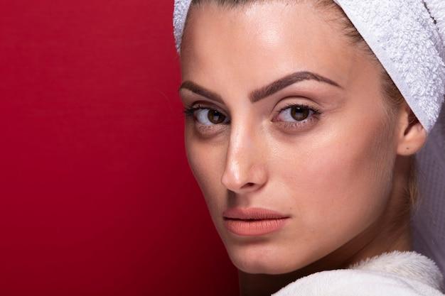 Portrait of a woman with a towel wrapped around her head against a red background, showcasing clear skin and subtle makeup